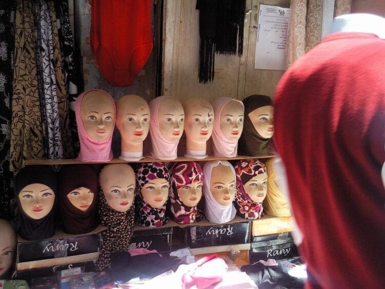 A woman wearing a hijab looks at a store shelf of mannequin heads displaying hijabs.