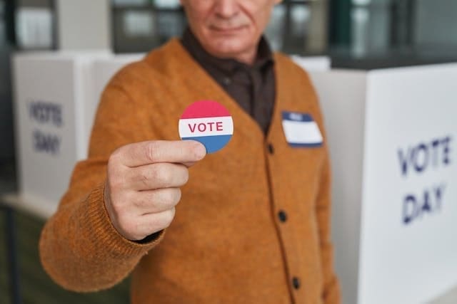 Voters against their best interests. A man in a brown sweater holds up a round sticker that says VOTE